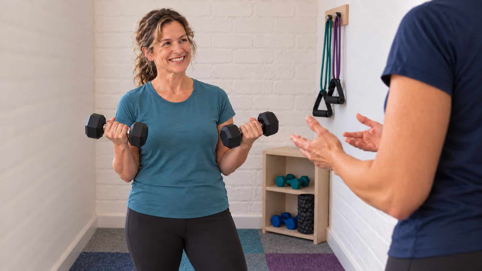 A smiling woman lifting dumbbells with guidance from a trainer